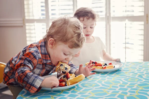 Zwei glückliche Kinder Zwillinge Junge Mädchen beim Frühstück Waffeln mit Früchten sitzen am Tisch in der sonnigen Küche frühmorgens mit Sonnenlicht von hinten — Stockfoto