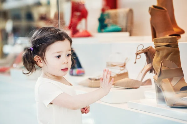 Closeup portrait of cute adorable sad upset white Caucasian toddler girl child with dark brown eyes and curly pig-tails hair in white light dress tshirt in mall looking at shoes in shop store window