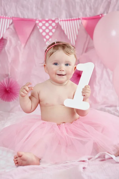 Portrait of cute adorable Caucasian baby girl with blue eyes in pink tutu skirt celebrating her first birthday with gourmet cake and balloons looking in camera, cake smash first year concept