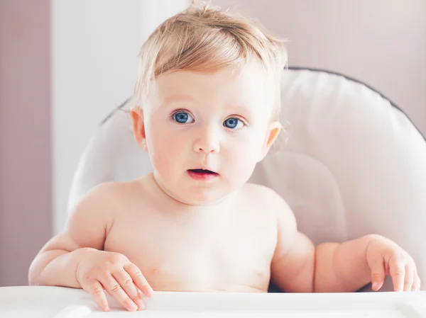 Closeup portrait of cute adorable funny blonde Caucasian smiling laughing baby boy girl with blue eyes with emotional face expression sitting in high chair in kitchen looking away