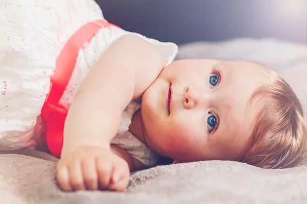 Portrait de mignon adorable blonde caucasien souriant bébé fille aux yeux bleus en robe blanche avec arc rouge couché sur le lit en regardant vers le haut rêvant, conte de fées lumière du soleil fuite rayon faisceau d'en haut derrière — Photo