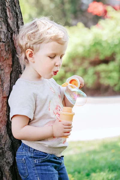 Porträtt av söt rolig liten blond kaukasiska barn flicka småbarn står i gröna skogen fältet ängen blåser Såpbubblor, ljusa sommardag, sommarnöje — Stockfoto