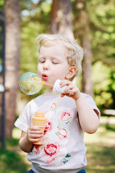 Porträtt av söt rolig liten blond kaukasiska barn flicka småbarn står i gröna skogen fältet ängen blåser Såpbubblor, ljusa sommardag, sommarnöje — Stockfoto