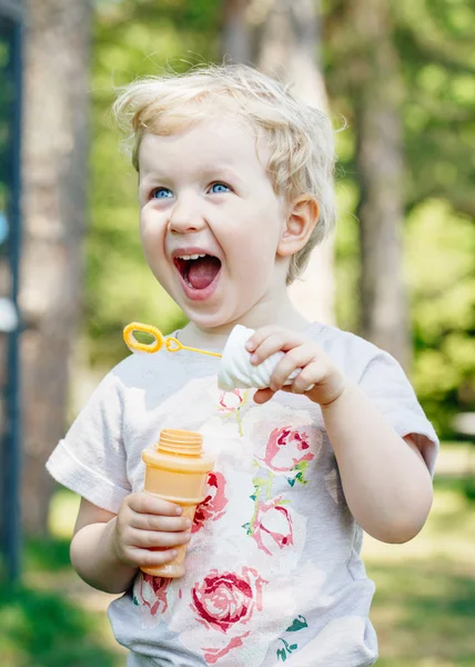 Porträtt av söt rolig liten blond kaukasiska barn flicka småbarn står i gröna skogen fältet ängen blåser Såpbubblor, ljusa sommardag, sommarnöje — Stockfoto