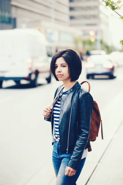 Πορτρέτο του όμορφη νεαρή γυναίκα καυκάσιος latino κορίτσι με σκούρα καστανά μάτια και μικρή σκούρα μαλλιά στο μπλε τζιν, δερμάτινο μπουφάν ποδηλάτης με σακίδιο που στέκεται στο δρόμο έξω, αστική πόλη προβολή φόντου — Φωτογραφία Αρχείου