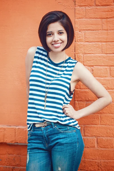 Retrato de bela sorridente jovem hipster latino hispânico menina mulher com cabelo curto bob, em jeans azuis, tshirt listrada, inclinando-se na parede de tijolo vermelho na cidade olhando para a câmera, tonificado com filtros Instagram — Fotografia de Stock