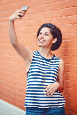 Çizgili tshirt ve mavi jeans ile karanlık kısa uzun bob saç kesimi dışında kırmızı tuğla duvar selfie fotoğraf cep telefonu ile yapma City yakınındaki güzel genç latin Latin kız kadın portresi