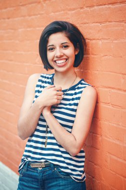 Portrait of beautiful smiling young hipster latin hispanic girl woman with short hair bob, in blue jeans, striped tshirt, leaning on red brick wall in city looking in camera, toned with Instagram filters clipart