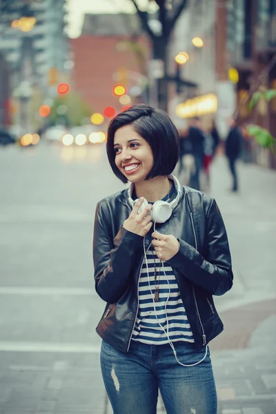 Πορτρέτο του όμορφη Ισπανόφωνος Λατινική κορίτσι γυναίκα κοντά μαύρα μαλλιά από δέρμα σακάκι με ακουστικά έξω στο δρόμο της πόλης το βράδυ βράδυ χαμογελώντας γελώντας ψάχνει μακριά, πορτραίτο έννοια τρόπου ζωής — Φωτογραφία Αρχείου