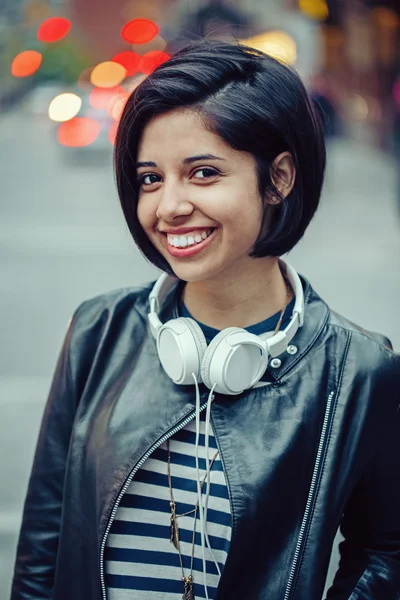 Portrait of beautiful Hispanic latin girl woman short black hair in leather jacket with headphones outside in evening night city street smiling laughing looking in camera, lifestyle portrait concept