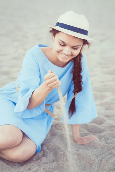 Retrato de mujer morena blanca sonriente caucásica con piel bronceada en vestido azul y sombrero de paja sentado en la playa de arena, puesta de sol en el día de verano, concepto de estilo de vida — Foto de Stock