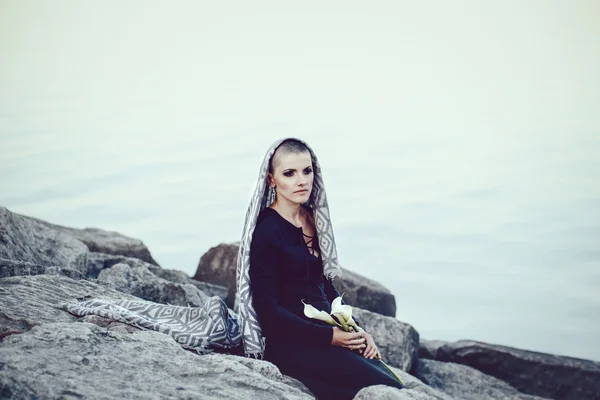 Portrait of sad beautiful Caucasian white young bald girl woman with shaved hair head covered with scarf shawl looking away, sitting by water on rocks holding lily flowers, toned with Instagram filters — Stock Photo, Image