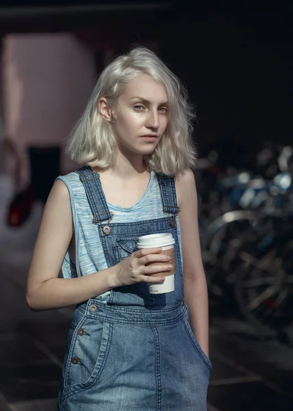 Portrait of beautiful Caucasian teenage young blonde alternative model girl woman in blue tshirt, jeans romper looking in camera holding cup of coffee, toned with Instagram filters