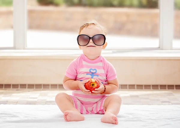 Portrait of funny cute white Caucasian smiling baby girl wearing large sunglasses sitting on floor in swimming pool by large window looking in camera, eating apple — Stock Photo, Image