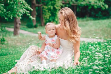 İki kişilik grup, beyaz beyaz anne ve beyaz elbiseli kız bebek çocuk dışarıda yeşil yazlık parkta oynarken, kameraya bakıyor, yaşam tarzı mutlu çocukluk konsepti