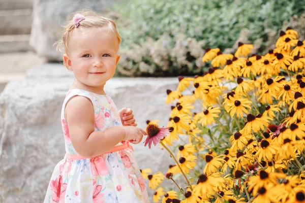 Lindo adorable blanco caucásico bebé niña en vestido blanco de pie entre flores amarillas fuera en jardín parque buscando en cámara, estilo de vida feliz concepto de la infancia — Foto de Stock