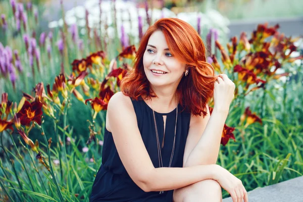 Portrait rapproché de souriante femme caucasienne blanche d'âge moyen aux cheveux roux bouclés ondulés en robe noire regardant loin dans le jardin du parc parmi les fleurs, concept de mode de vie beauté — Photo