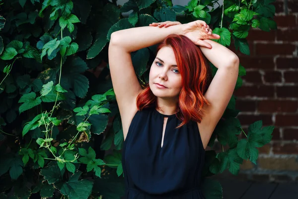 Closeup portrait of relaxing sexy middle aged white caucasian woman with waved red hair in black dress looking in camera outside in green plant leaves with hands up, beauty fashion