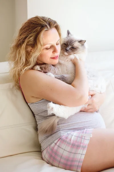 Retrato de mujer embarazada rubia blanca joven caucásica con pelo largo en camiseta y pantalones cortos relajándose en casa en el sofá sosteniendo gato, concepto de estilo de vida de maternidad saludable — Foto de Stock