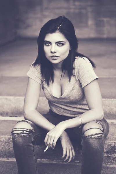 Toned black and white portrait of white Caucasian beautiful young sexy brunette woman in ripped jeans and t-shirt holding smoking cigarette looking in camera, lifestyle concept — Stock Photo, Image