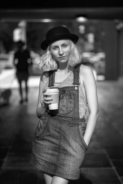 Zwart-wit portret van Kaukasische teenage jonge blonde alternatief model meisje vrouw in tshirt, jeans romper kijken in de camera bedrijf kopje koffie — Stockfoto