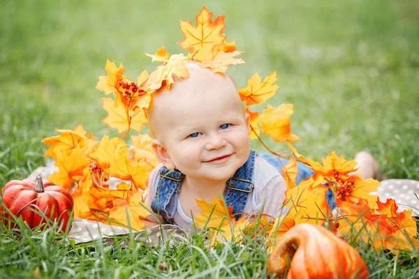 Portret van leuke grappige schattige blonde Kaukasische babyjongen met blauwe ogen in t-shirt en spijkerbroek romper liggen op gras veld weide, gele herfst herfstbladeren pompoenen. Halloween, Thanksgiving. — Stockfoto