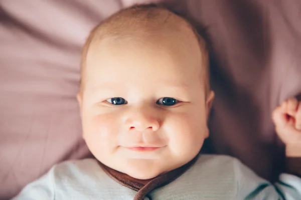 Closeup portrait of cute adorable funny white Caucasian blond little baby boy newborn with blue grey eyes lying on bed looking in camera smiling, lifestyle candid, real life