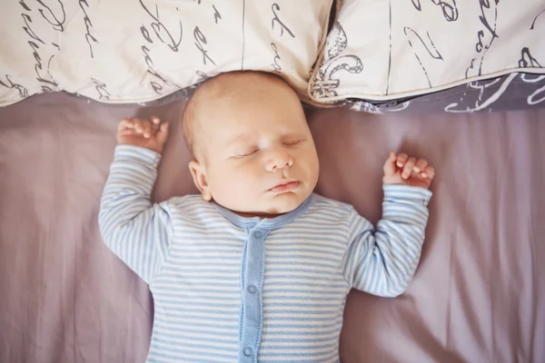 Ritratto di dormire divertente bianco caucasico biondo bambino neonato in abiti blu sdraiato sul letto con le mani in alto, stile di vita candido concetto reale — Foto Stock