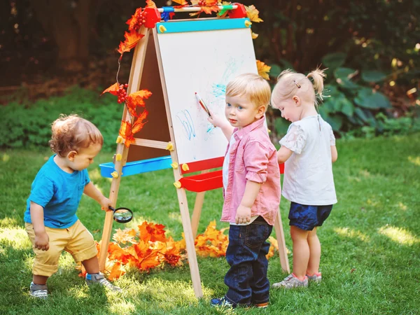 Group of three white Caucasian toddler children kids boys and girl standing outside in summer autumn park by drawing easel with markers, playing studying learning, back to school