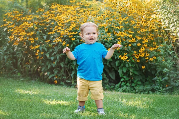 Porträtt av söt bedårande skrattande leende vit kaukasiska baby pojke barn stående bland gula blommor utanför i trädgården park tittar bort, lycklig barndom livsstilskoncept — Stockfoto