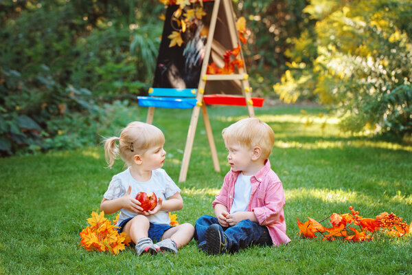 Group of two white Caucasian toddler children kids boy and girl sitting outside in summer autumn park by drawing easel looking at each other, playing eating apple, back to school