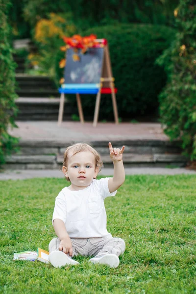 Portrait d'enfant tout-petit caucasien blanc drôle enfant garçon assis sur le sol d'herbe à l'extérieur dans le parc d'automne d'été en dessinant chevalet avec des livres montrant signe de salut en métal de roche, retour à l'école — Photo