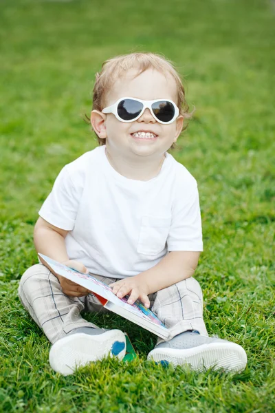 Retrato de engraçado bonito adorável sorrindo rindo branco caucasiano criança menino com cabelo loiro em t-shirt branca e óculos de sol sentado na grama verde segurando um livro . — Fotografia de Stock