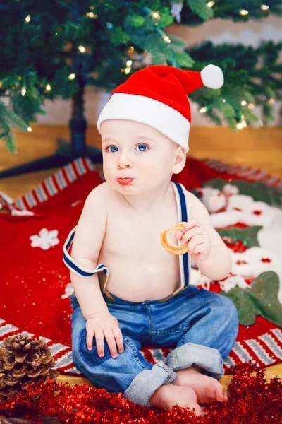 Lifestyle portrait of happy funny white Caucasian baby boy in jeans and New Year Christmas Santa hat sitting on floor eating cookie indoor at home looking in camera — Stock Photo, Image