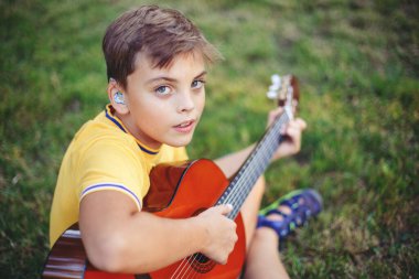Dışarıda gitar çalan reşit olmayan bir çocuğu duymakta zorlanıyorum. Kulağında işitme cihazı olan, müzik çalan ve parkta şarkı söyleyen bir çocuk. Çocuklar için hobi sanatı aktivitesi. Otantik çocukluk anı. 