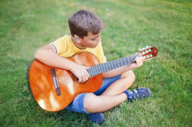 Dışarıda gitar çalan reşit olmayan bir çocuğu duymakta zorlanıyorum. Kulağında işitme cihazı olan, müzik çalan ve parkta şarkı söyleyen bir çocuk. Çocuklar için hobi sanatı aktivitesi. Otantik çocukluk anı. 