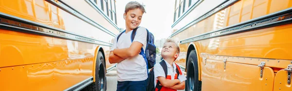Happy Caucasian brothers students near yellow school bus. Smiling kids going back to school in September. Education system and learning. Support and friendship. Web banner header.