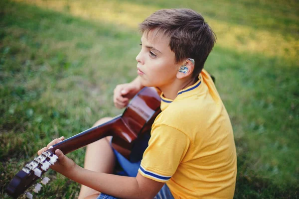 Dışarıda gitar çalan reşit olmayan bir çocuğu duymakta zorlanıyorum. Kulağında işitme cihazı olan, müzik çalan ve parkta şarkı söyleyen bir çocuk. Çocuklar için hobi sanatı aktivitesi. Otantik çocukluk anı. 
