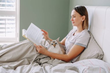 Güzel, mutlu, orta yaşlı bir kadın evde oturmuş kitap okuyor. Kedi evcil hayvanı olan genç bir kadın, elinde kitap tutuyor. Rahatla, evde dinlenme zamanı. Yavaş yaşam ve hayatın basit zevkleri.