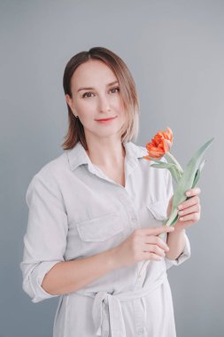 Mutlu gülümseyen orta yaşlı beyaz bir kadının portresi kırmızı lale çiçeği tutuyor. Gri ışıklı arka planda kısa saçlı gerçek bir kadın. Doğal güzellik görünüşü. Anneler günü tatili.