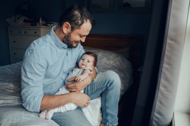 Orta yaşlı beyaz bir baba kucağında bebek sallıyor. Evde elinde çocuğuyla yatakta oturan yorgun bir ebeveyn. Otantik yaşam tarzı aile anı. Bekar baba ev hayatı. 