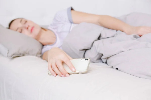 Woman lying in bed in morning with smartphone. Young woman addictive to online Internet. Person resting sleeping with phone in hand. Digital online Internet addiction.