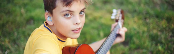 Hard of hearing preteen boy playing guitar outdoor. Child with hearing aids in ears playing music and singing song in park. Hobby art activity for children kids. Web banner header.