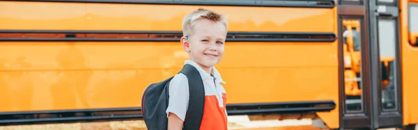 Happy Caucasian boy student with backpack near yellow bus on first September day. Education and back to school in autumn fall. Child kid on schoolyard outdoor. Web banner header.