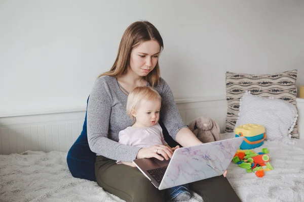 Young Caucasian mother with baby working on laptop from home. Workplace of freelance woman student with kid toddler. Stay at home single mom earning money at distant remote job online.