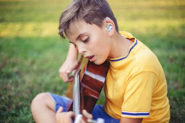Genç bir çocuğun gitar çalıp şarkı söylemesini duymakta zorlanıyorum. Kulağında işitme cihazı olan, müzik çalan ve parkta şarkı söyleyen bir çocuk. Çocuklar için hobi sanatı aktivitesi. Otantik çocukluk anı. 