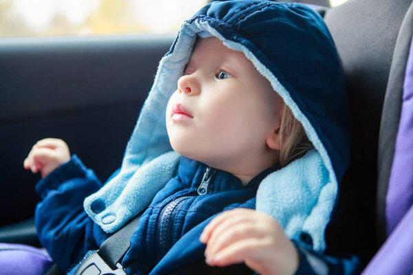 Cute Caucasian baby infant sitting in car seat. Adorable kid in outwear clothes in automobile vehicle carsit fastened with seatbelt. Care, safety and protection of children on roads.