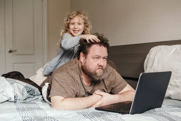 和孩子一起在家里工作 父亲背着女儿在卧室的笔记本电脑上工作 有趣而坦率的家庭时刻 食管动物检疫期间出现新的正常情况 — 图库照片