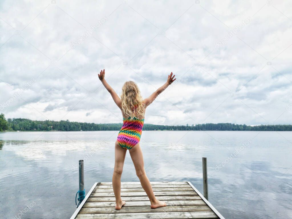 Blonde girl in swimsuit standing on wooden lake river dock with her hands raised up. Harmony with nature and happy healthy active childhood. Summer outdoor water activity. View from back. 
