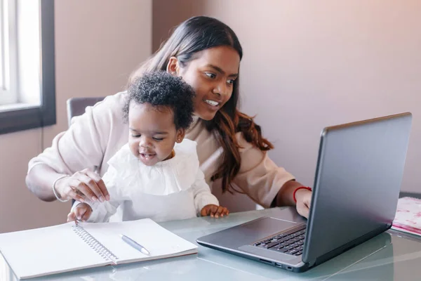 Mixed race Indian mother with African black baby working online from home on Internet. Workplace of freelancer woman with kid. Stay home single mom working distant job. New normal.